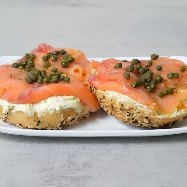 Two everything bagels with cream cheese, smoked salmon, and capers on a white plate.