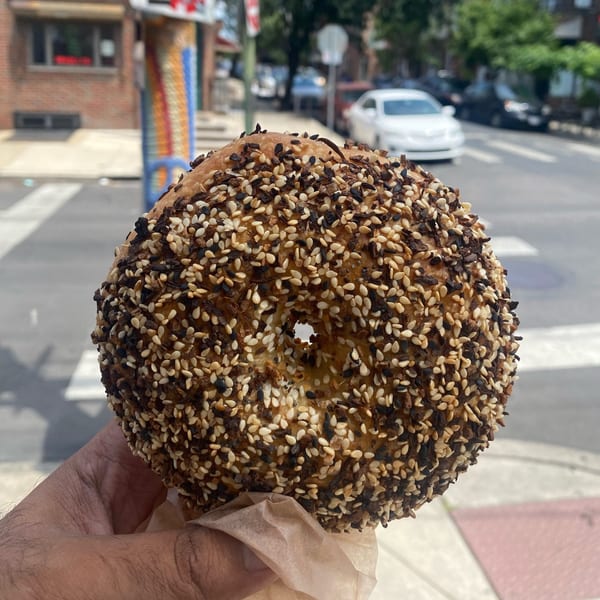 A hand holds an everything bagel from Slice & Schmear, with a street and cars in the background.