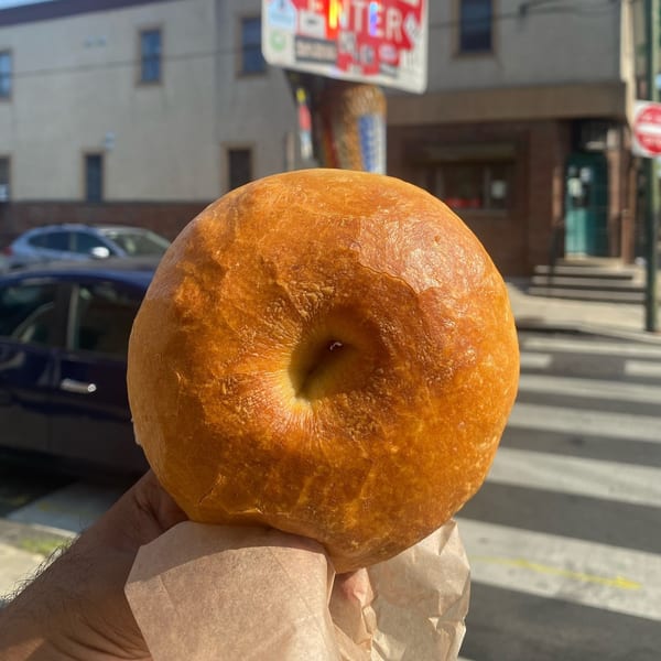 Hand holding a golden-brown bagel in a paper bag, with a city street in the background.