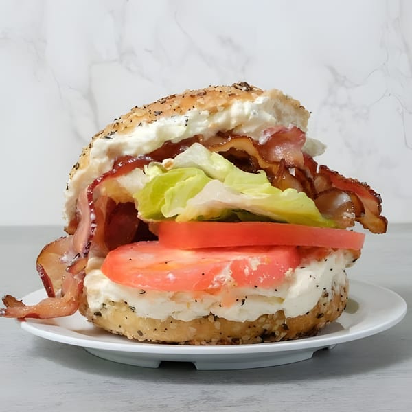 Everything bagel with cream cheese, bacon, lettuce, and sliced tomatoes on a white plate.