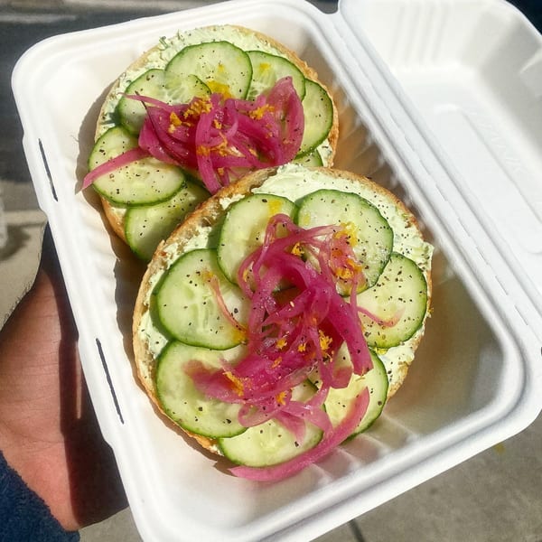 Two bagels with cream cheese, sliced cucumbers, pickled red onions, and black pepper in a to-go container.